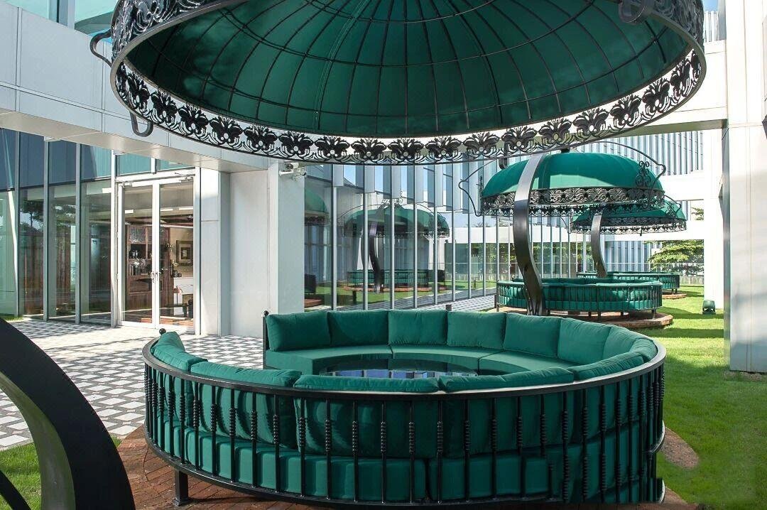 A modern, ornate black metal gazebo with a green fabric roof, featuring integrated circular green cushions for seating on a lawn.