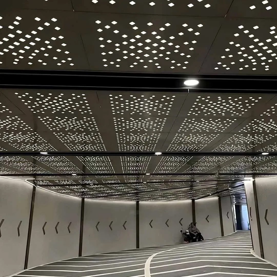 Square-pattern perforated stainless steel sheets installed as acoustic metal ceiling panels in a modern backlit corridor.
