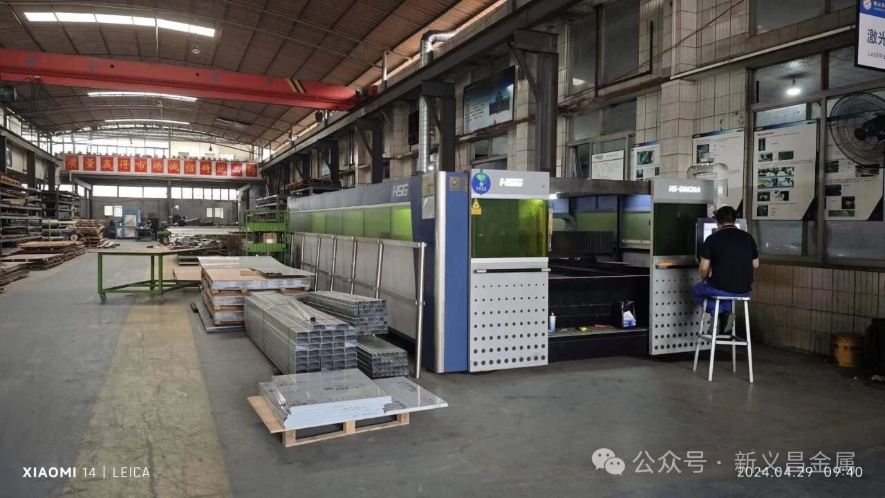 Workshop of XYC Metalworks, HSG fiber laser cutting machine. Technician operates the machine for stainless steel sheet cutting. Stainless steel plates and rectangular tubes stacked on site for custom architectural metal fabrication.